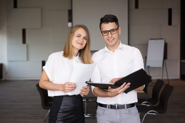 Portrait of happy male and female colleagues. Young Caucasian businessman wearing glasses standing with folder together with smiling female coworker in boardroom. Team concept